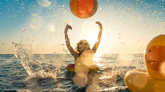 Ein glückliches Kind, das bei Sonnenuntergang im Wasser mit Wasserball und Badeente Spaß hat