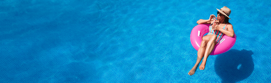 Junge Frau mit Sonnenbrille, Hut und Badeanzug in einem Pool, das den Sommer genießt und dabei einen Cocktail trinkt