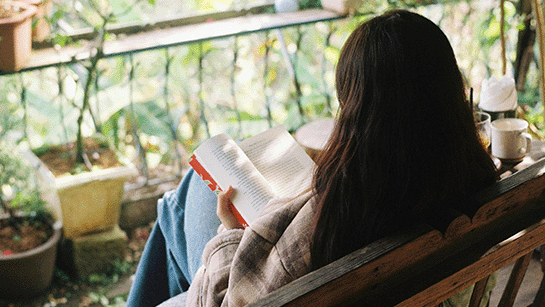Auf einer Bank sitzende Frau mit Buch auf dem Schoß, lesend auf Balkon