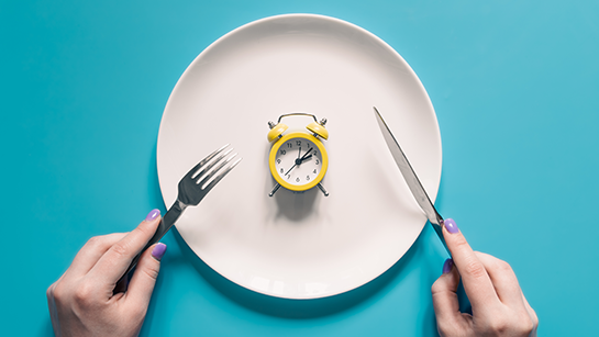 Hände halten Messer und Gabel über dem Wecker auf einem Teller auf blauem Hintergrund