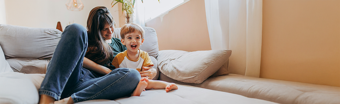 Titelbild Kategorie Gesundheitstipps Gesundheit: Eine Mutter sitzt mit Ihrem Kind auf dem Sofa und lacht.