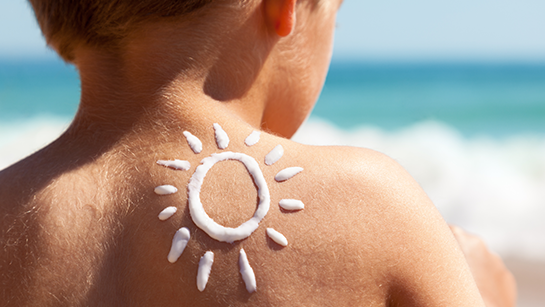Kleiner Junge in Rückenansicht mit einer Sonne aus Sonnencreme auf der Schulter schaut aufs Meer