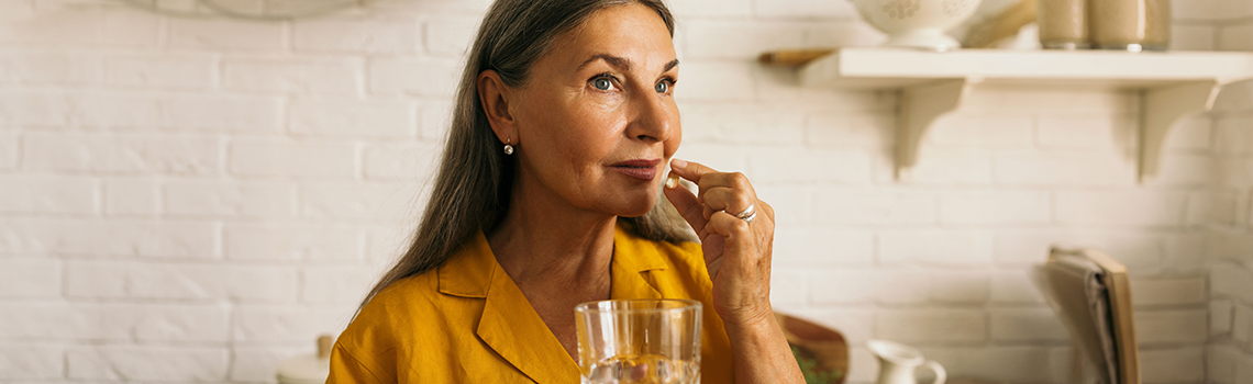 Frau mittleren Alters steht in der Küche mit einer Pille und einem Wasserglas in der Hand